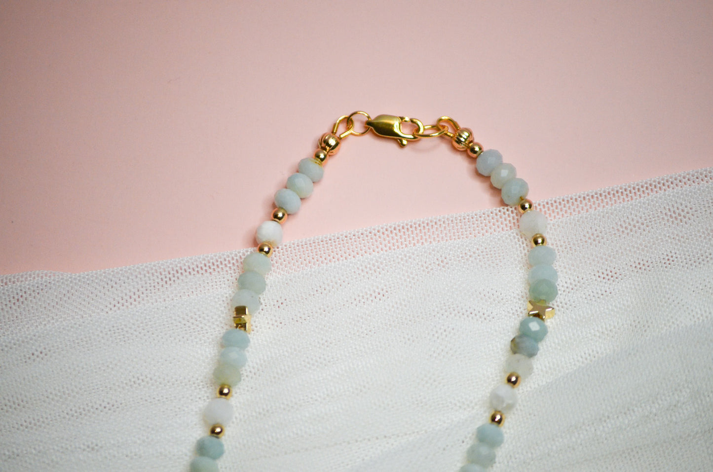 Necklace with gold and light blue beads, and a gold clasp on a pink and white fabric background
