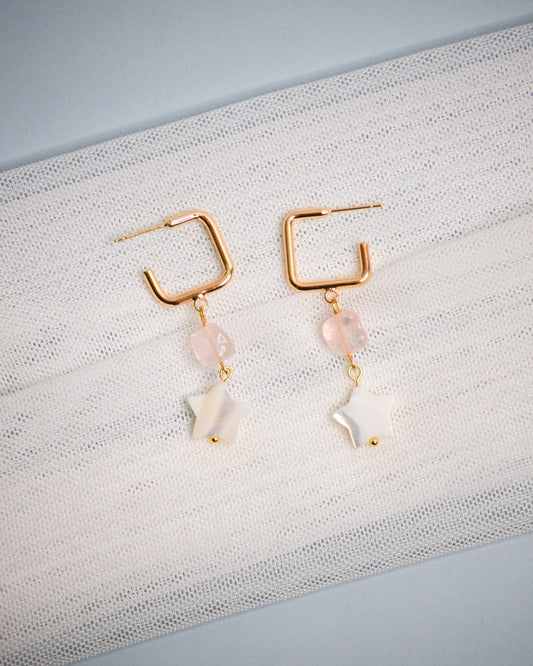 Pair of gold earrings with pink and white star-shaped beads on a textured fabric background