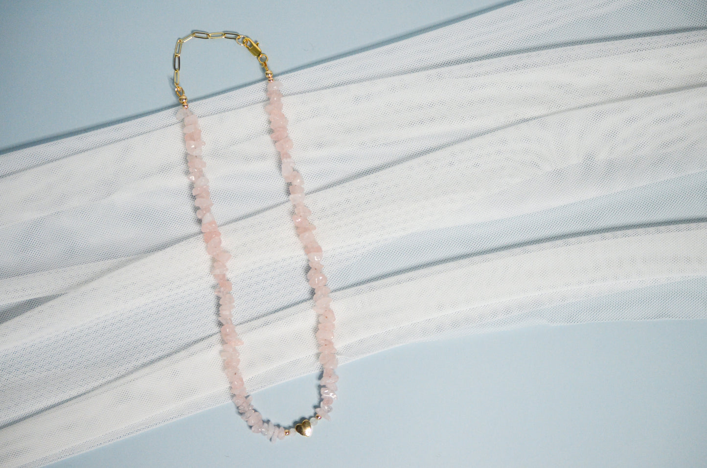Necklace with pink beads and a gold heart pendanton a light blue and white fabric background