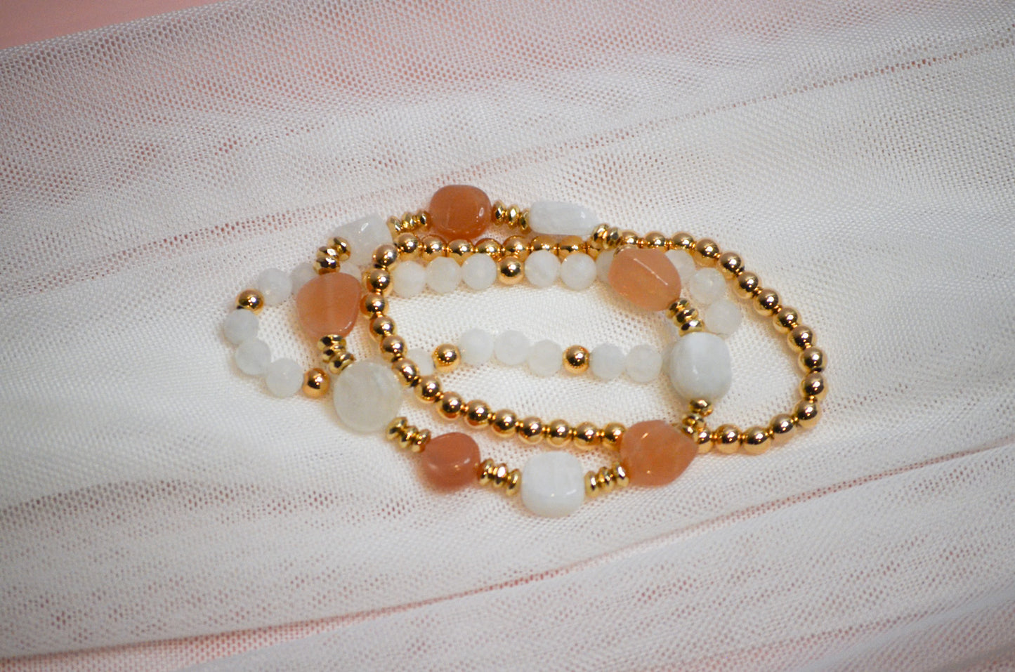 Set of three gold, pink, and white beaded bracelets on a textured fabric background
