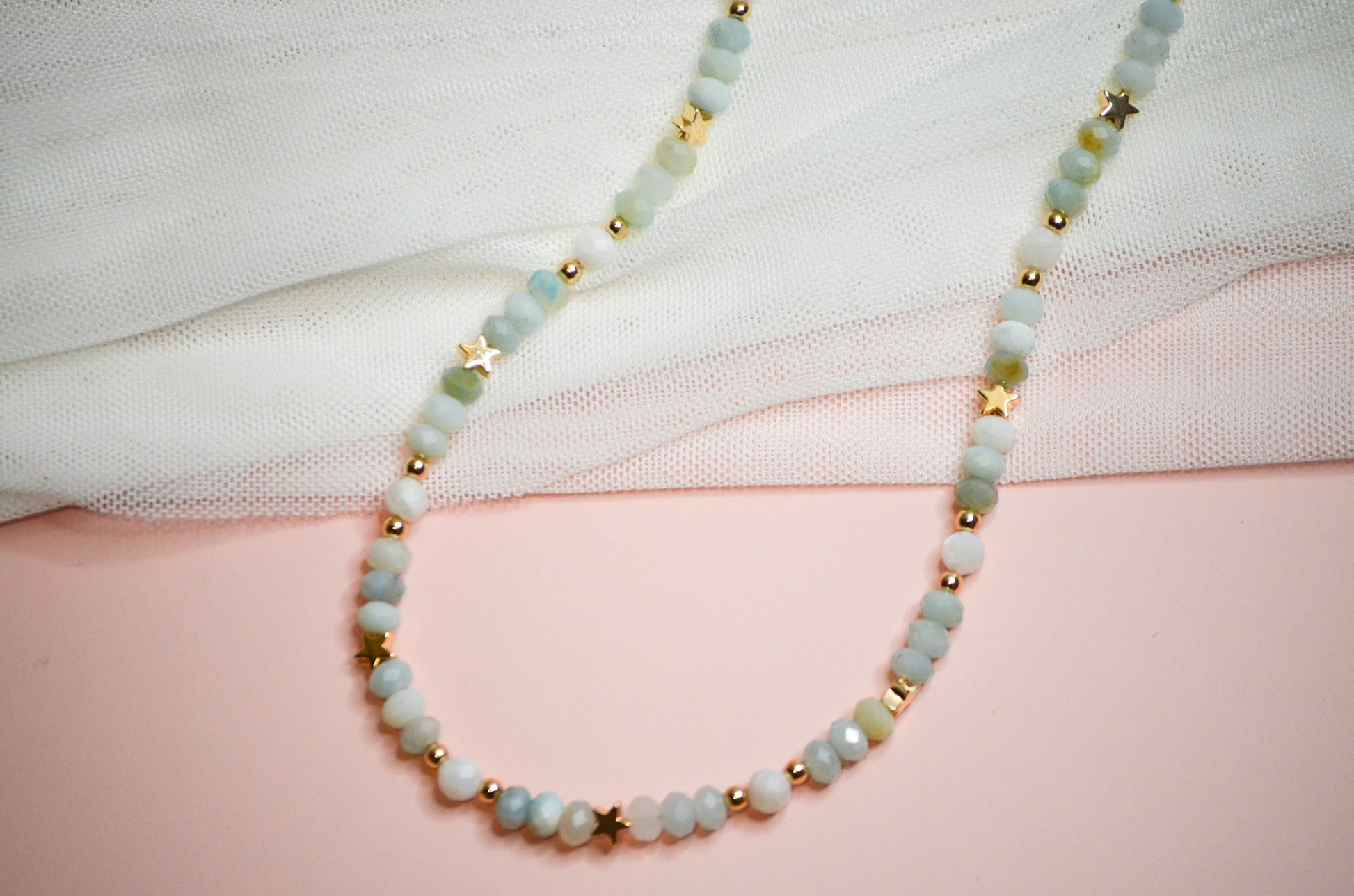 Necklace with light blue beads and gold star accents on a white and pink background