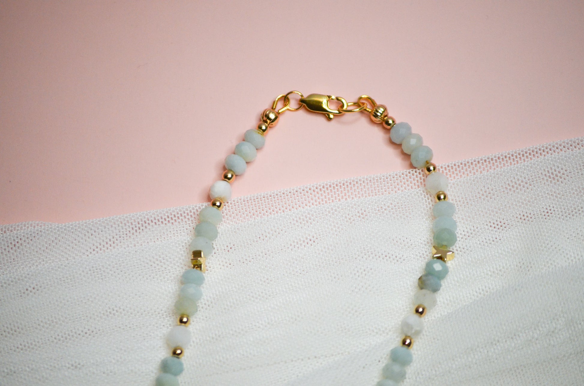 Necklace with gold and light blue beads, and a gold clasp on a pink and white fabric background