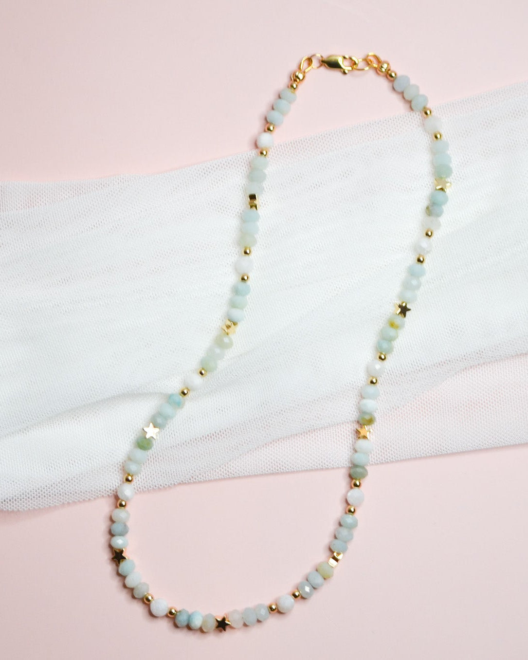 Necklace with light blue beads and gold star accents on a white and pink fabric background