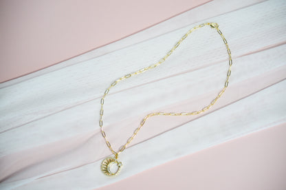 Gold necklace with a circle shaped pendant with a sun inside on a pink and white striped background.
