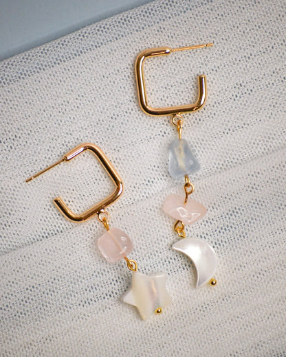 Pair of gold dangle earrings with pink, blue, and white beads on a textured surface. 