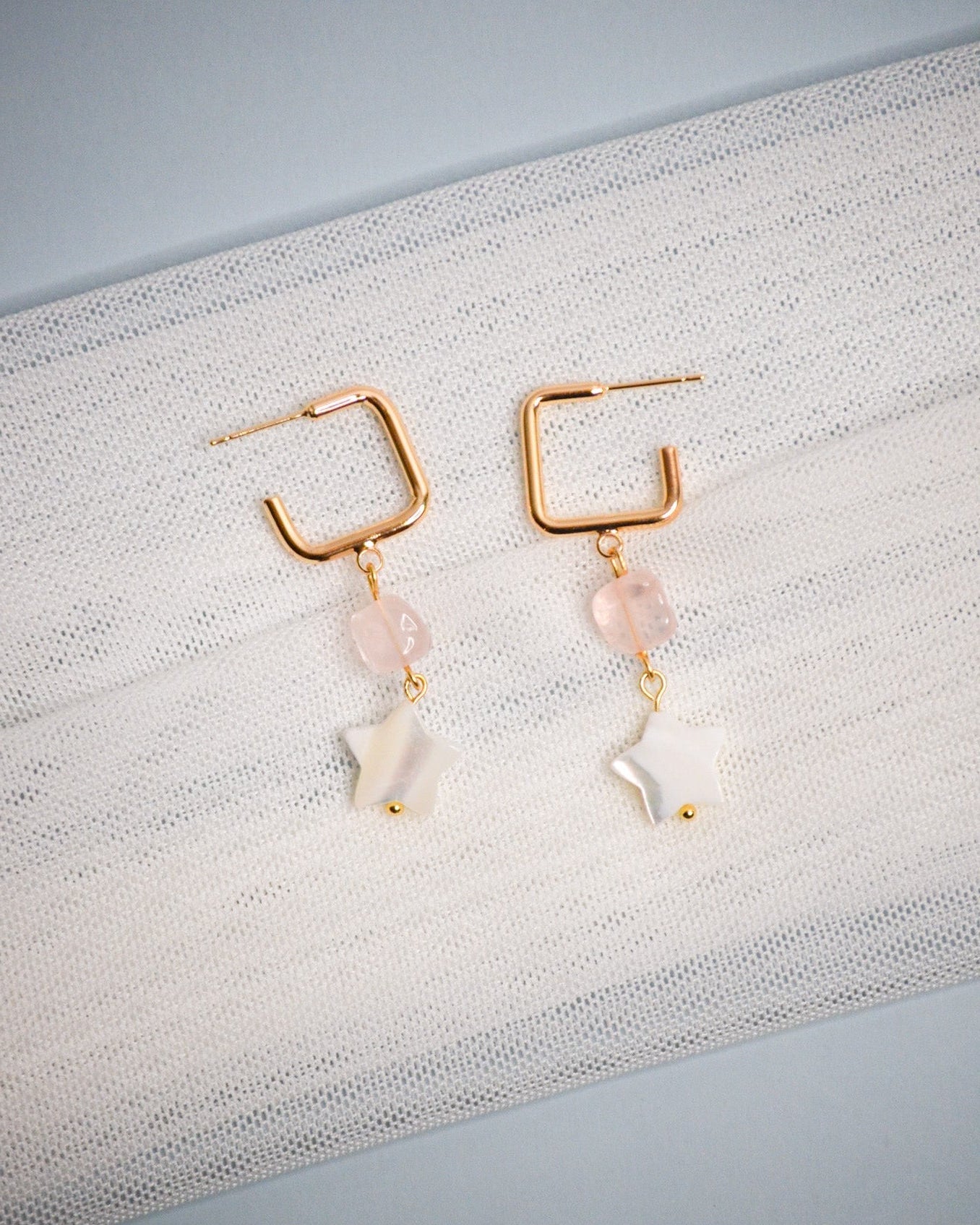 Pair of gold earrings with pink and white star-shaped beads on a textured fabric background