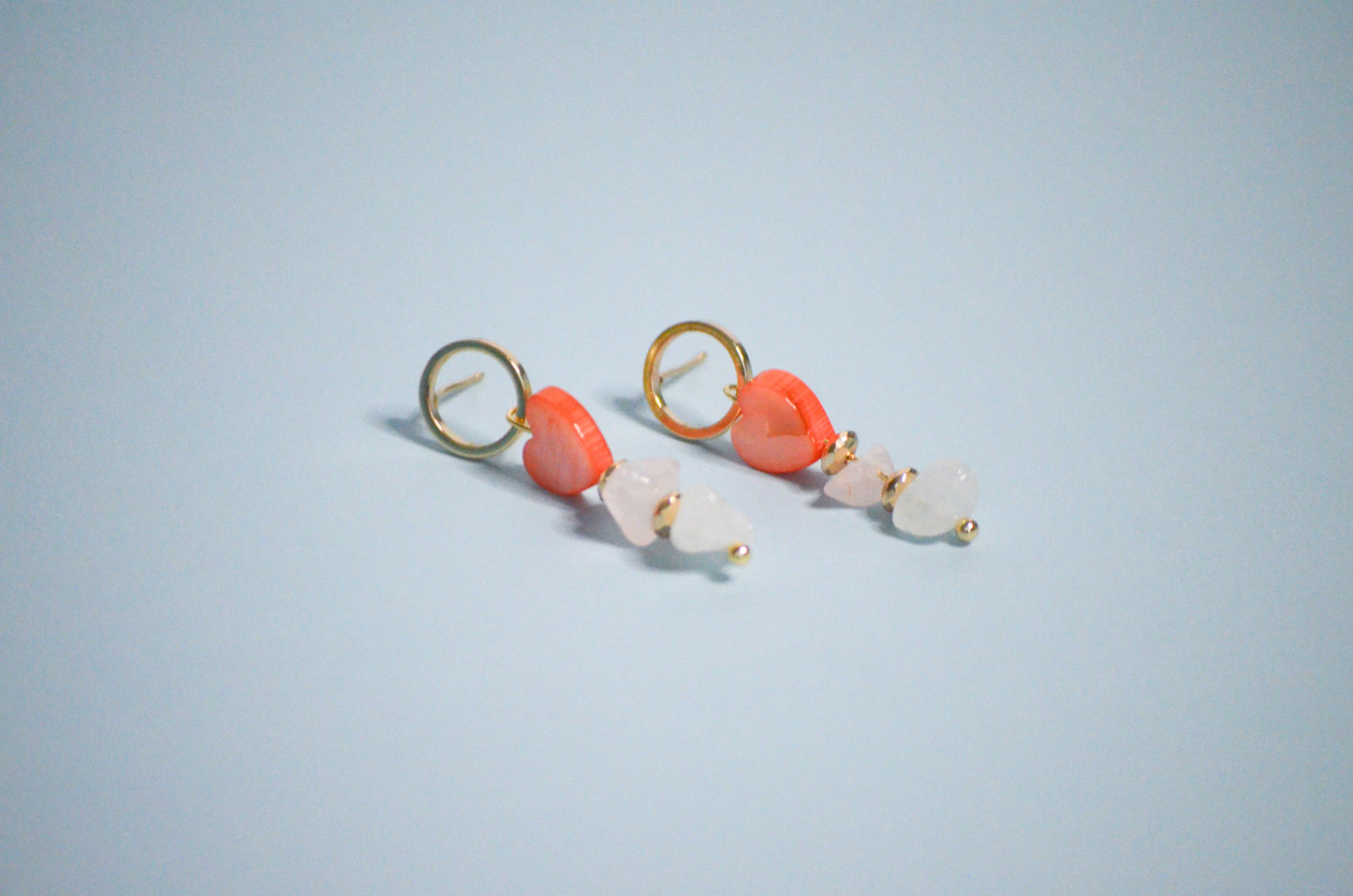 Pair of gold earrings with coral hearts,  clear, and gold beads on a light blue background