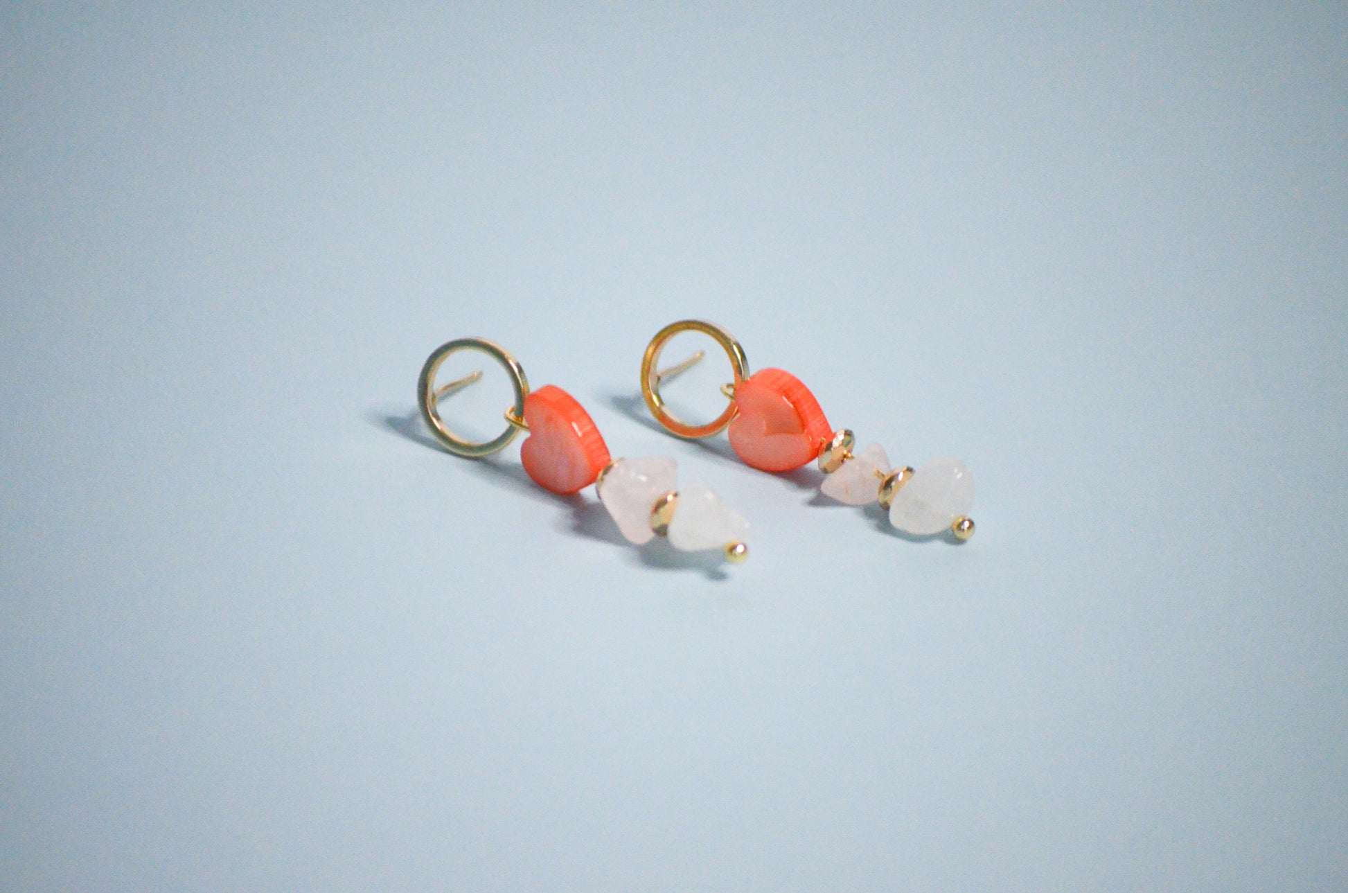 Pair of gold earrings with coral hearts,  clear, and gold beads on a light blue background