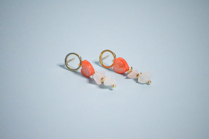 Pair of gold earrings with coral hearts,  clear, and gold beads on a light blue background