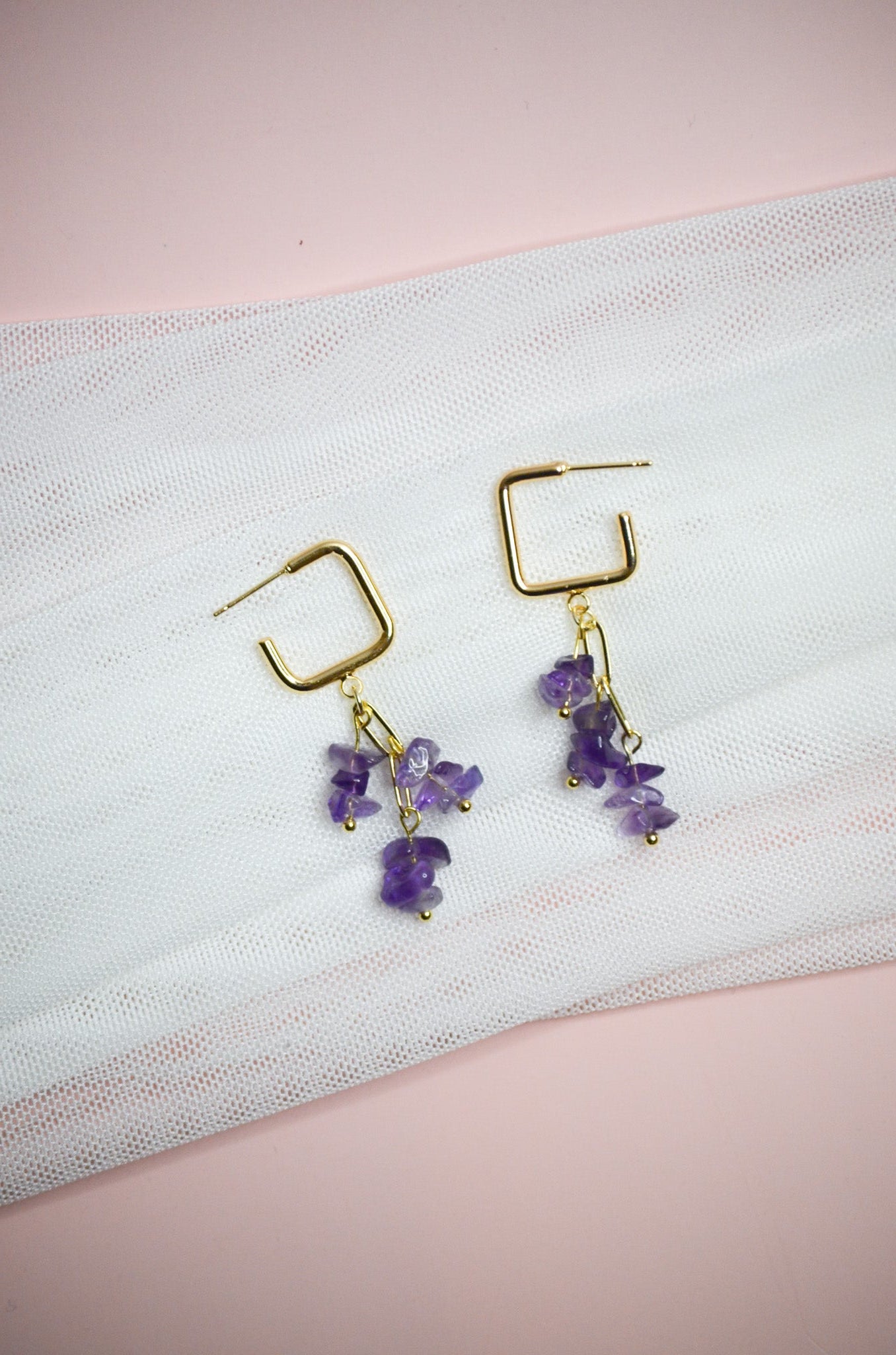 Gold square dangle earrings with purple stones on a white fabric background