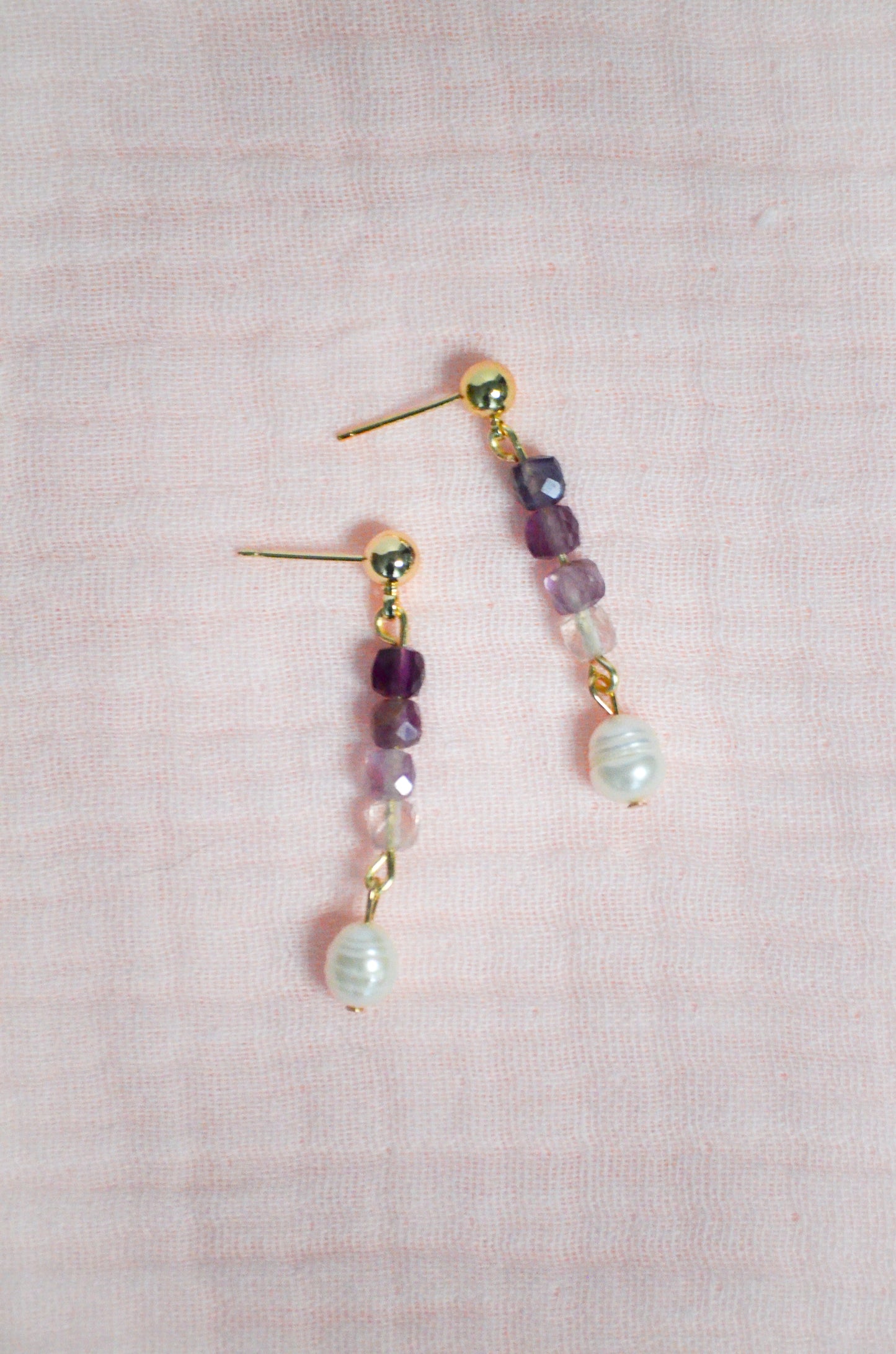 Pair of gold, purple gemstone, and pearl beaded earrings on a pink textured background