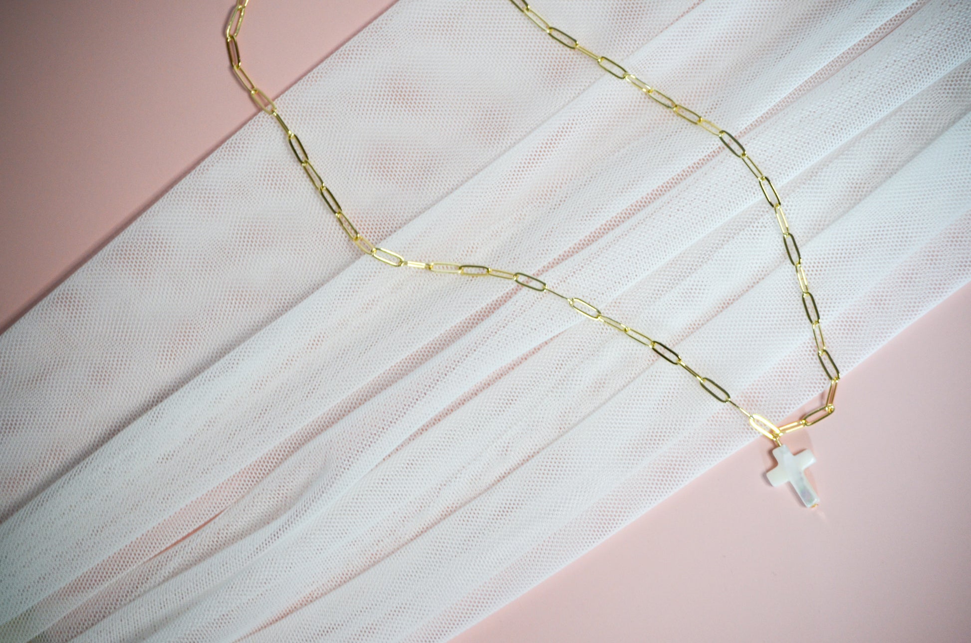 Gold paperclip chain necklace with a cross pendant on a pink and white textured background