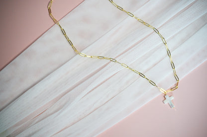 Gold paperclip chain necklace with a cross pendant on a pink and white textured background