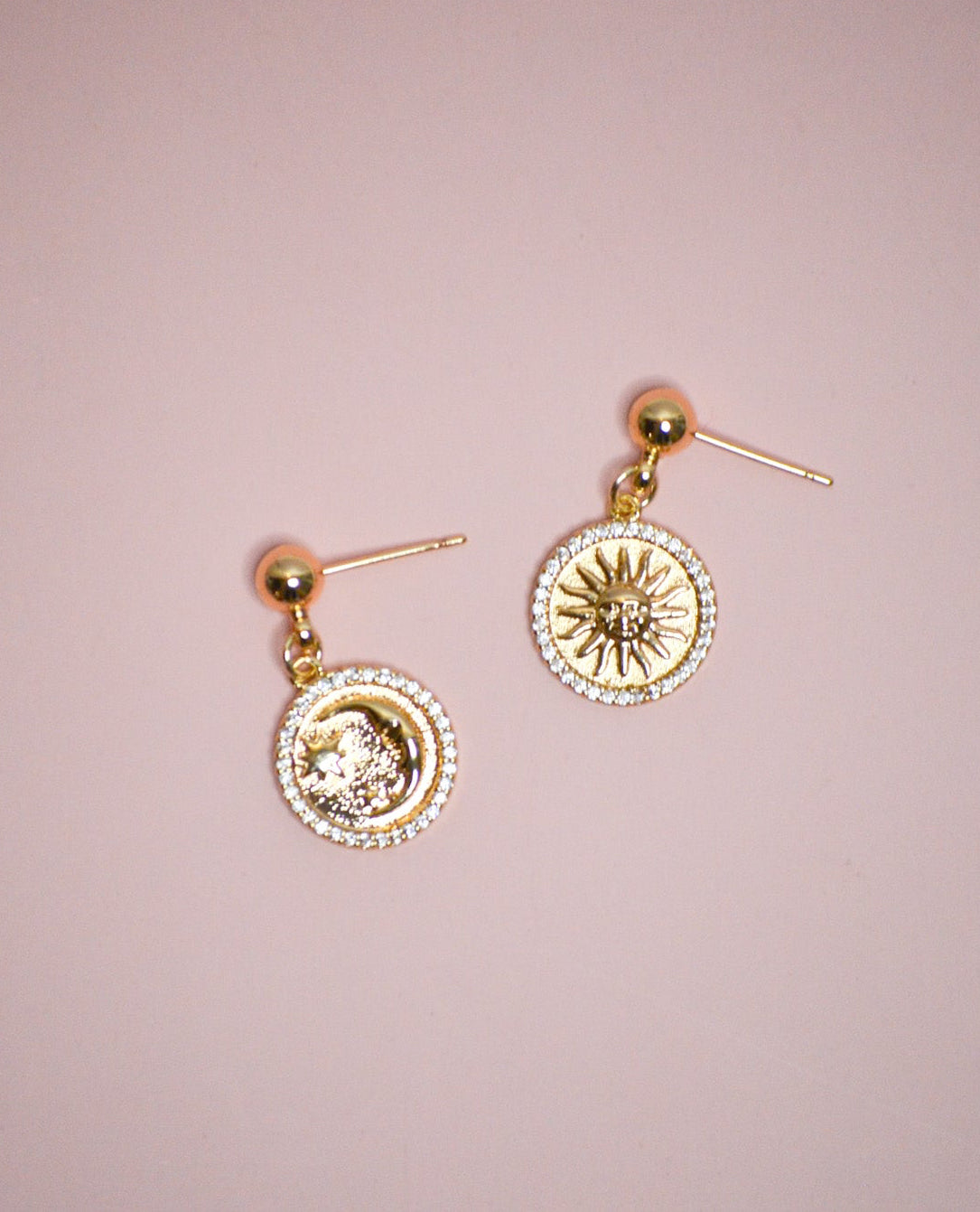 Gold circle dangle earrings with celestial designs on a pink background. 