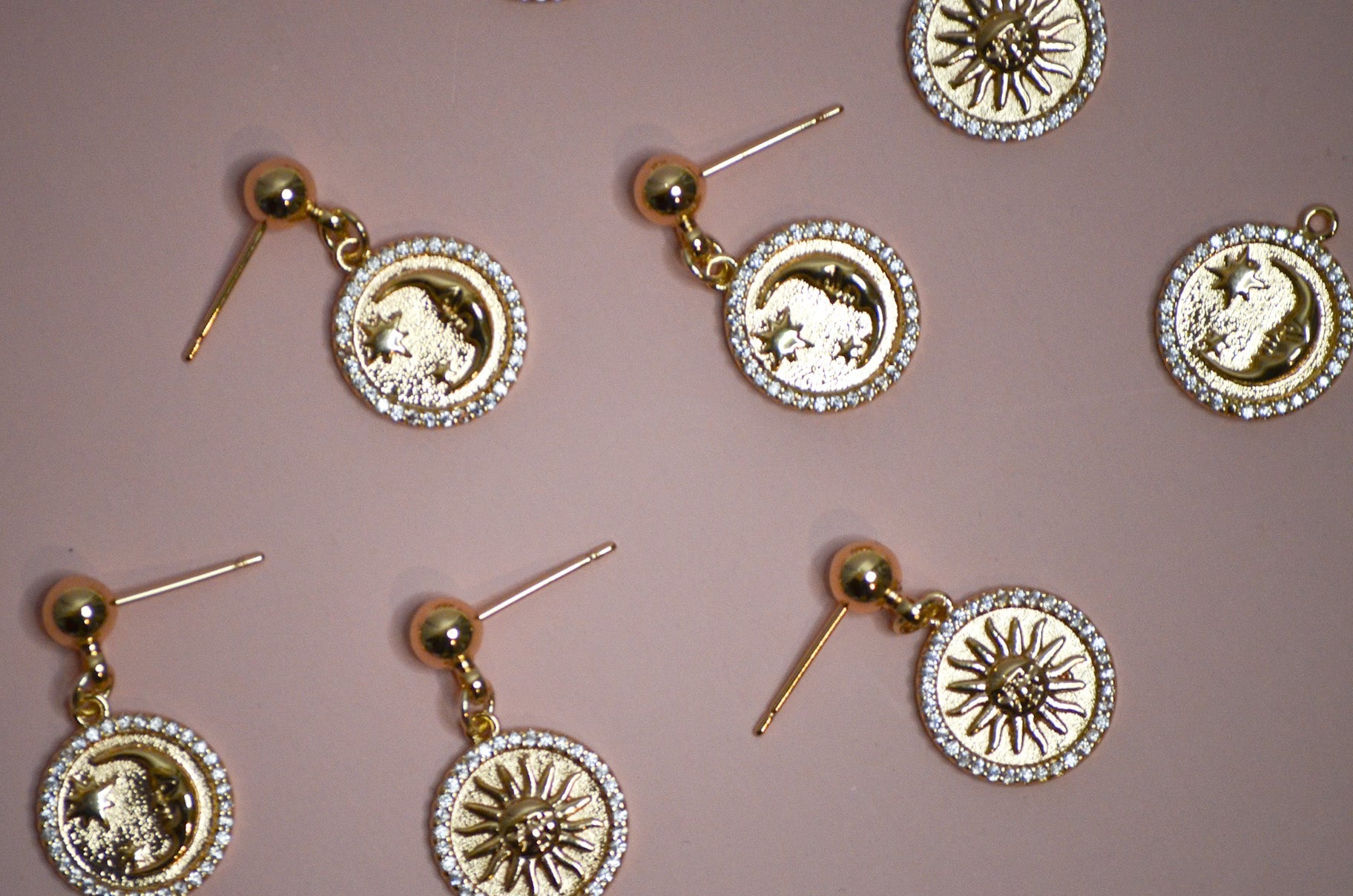 Seven gold dangle earrings with sun and moon designs on a pink background