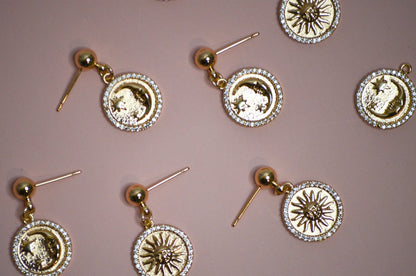 Seven gold dangle earrings with sun and moon designs on a pink background