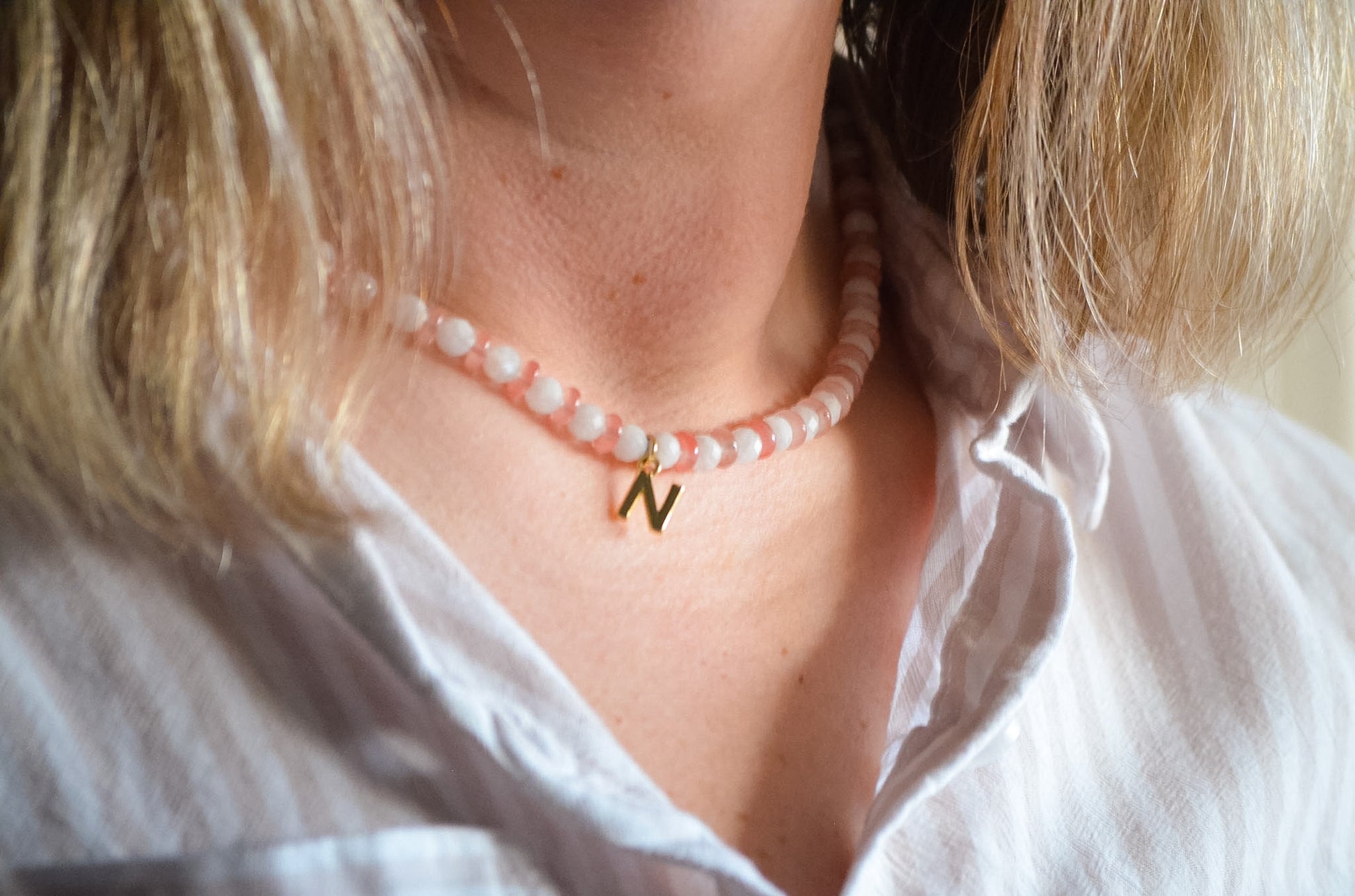 Woman with blonde hair wearing a pink and white beaded necklace with a gold .initial charm