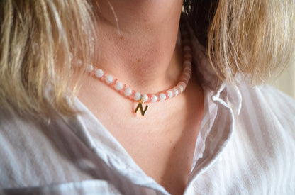 Woman with blonde hair wearing a pink and white beaded necklace with a gold .initial charm