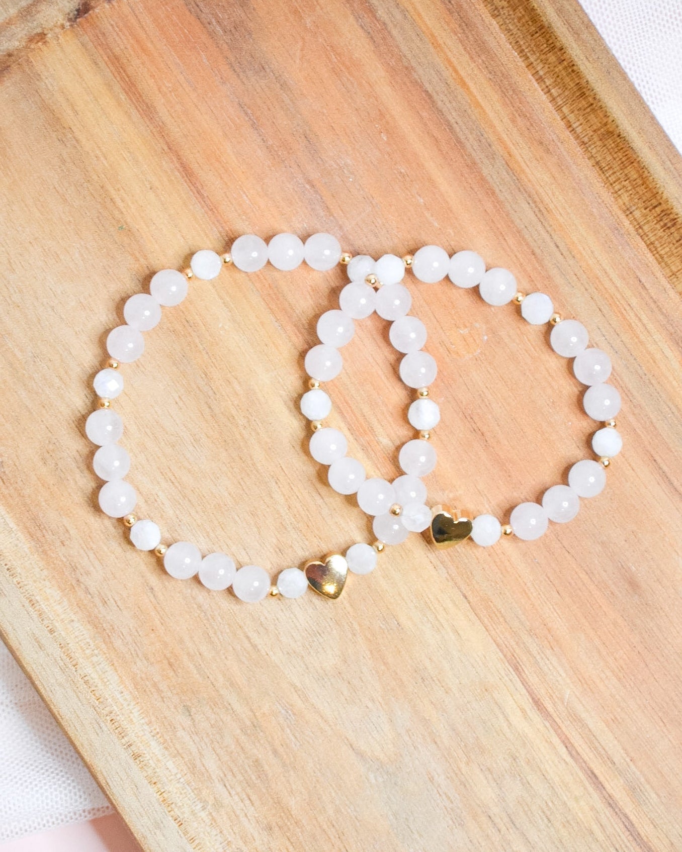 Two white beaded bracelets with a gold heart accent on a wooden surface