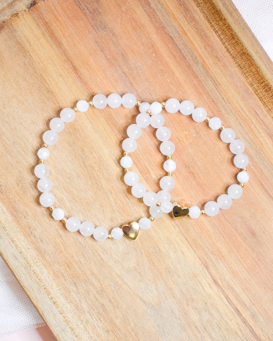 Two white beaded bracelets with a gold heart accent on a wooden surface