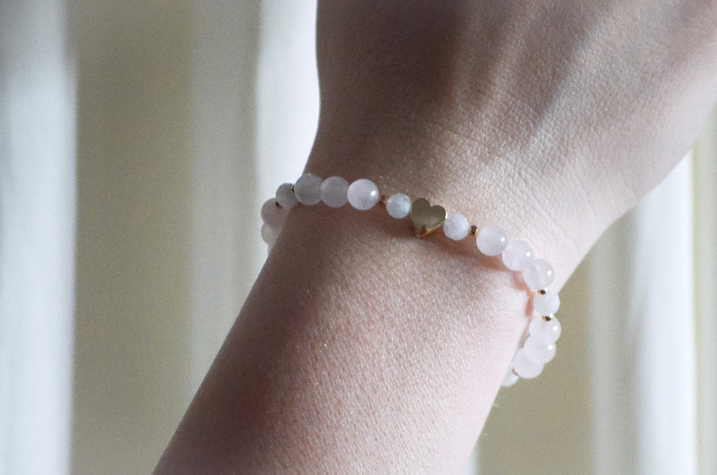 Close-up of a wrist wearing a bracelet with white beads and a gold heart on a neutral background