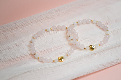 Two white beaded bracelets with gold heart accents on a sheer fabric background