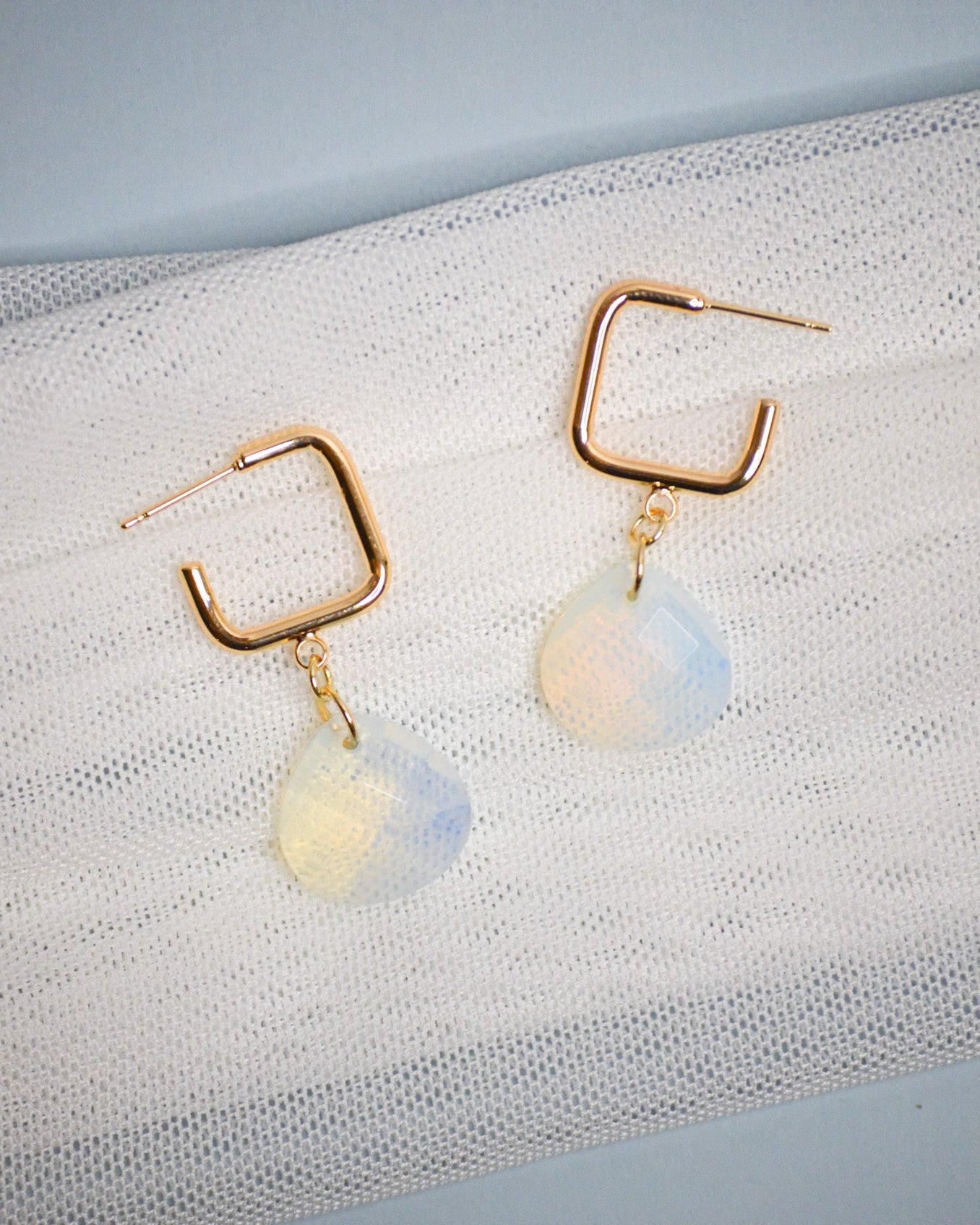 Gold square dangle hoop earrings with opalite stones on a textured white surface
