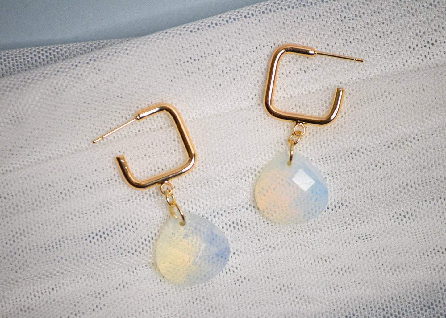 Gold square hoop earrings with teardrop shaped iridescent gemstones on a textured white fabric background