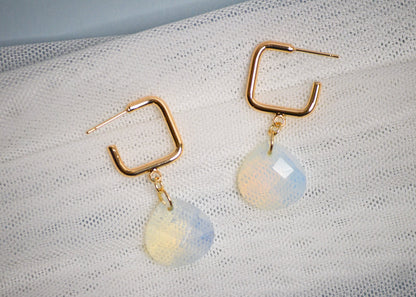 Gold square hoop earrings with teardrop shaped iridescent gemstones on a textured white fabric background