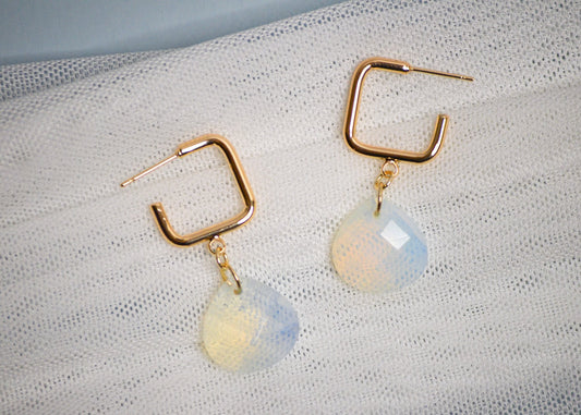 Gold square hoop earrings with teardrop shaped iridescent gemstones on a textured white fabric background