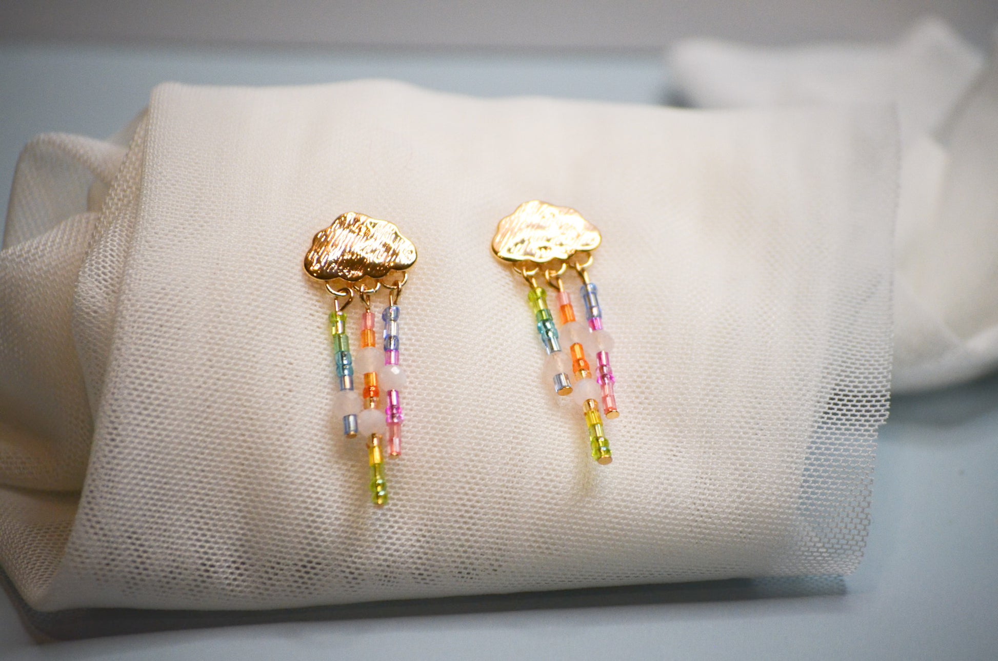 Gold cloud-shaped earrings with colorful rainbow raining beads on a white cushion
