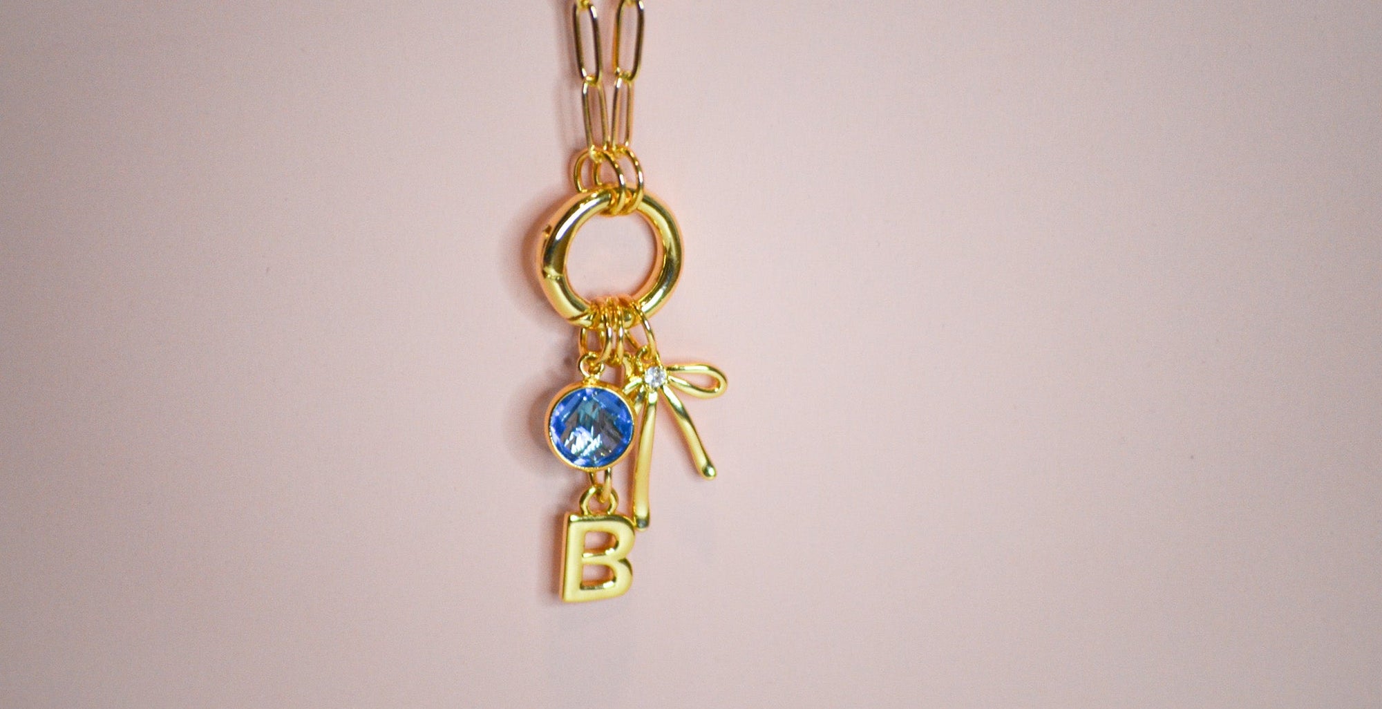 Gold necklace with three charms: blue gemstone, letter 'B', and a bow on a pink background