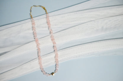 Necklace with pink beads and a gold heart pendanton a light blue and white fabric background