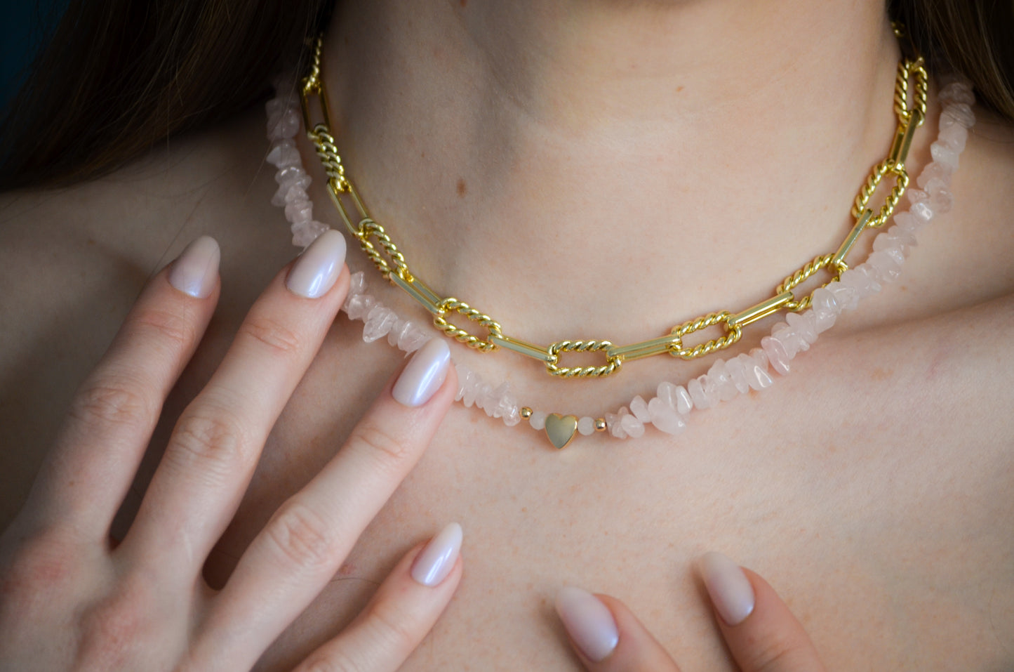 Gold chain necklace with pink stones and gold heart beads worn by a woman with light pink nails