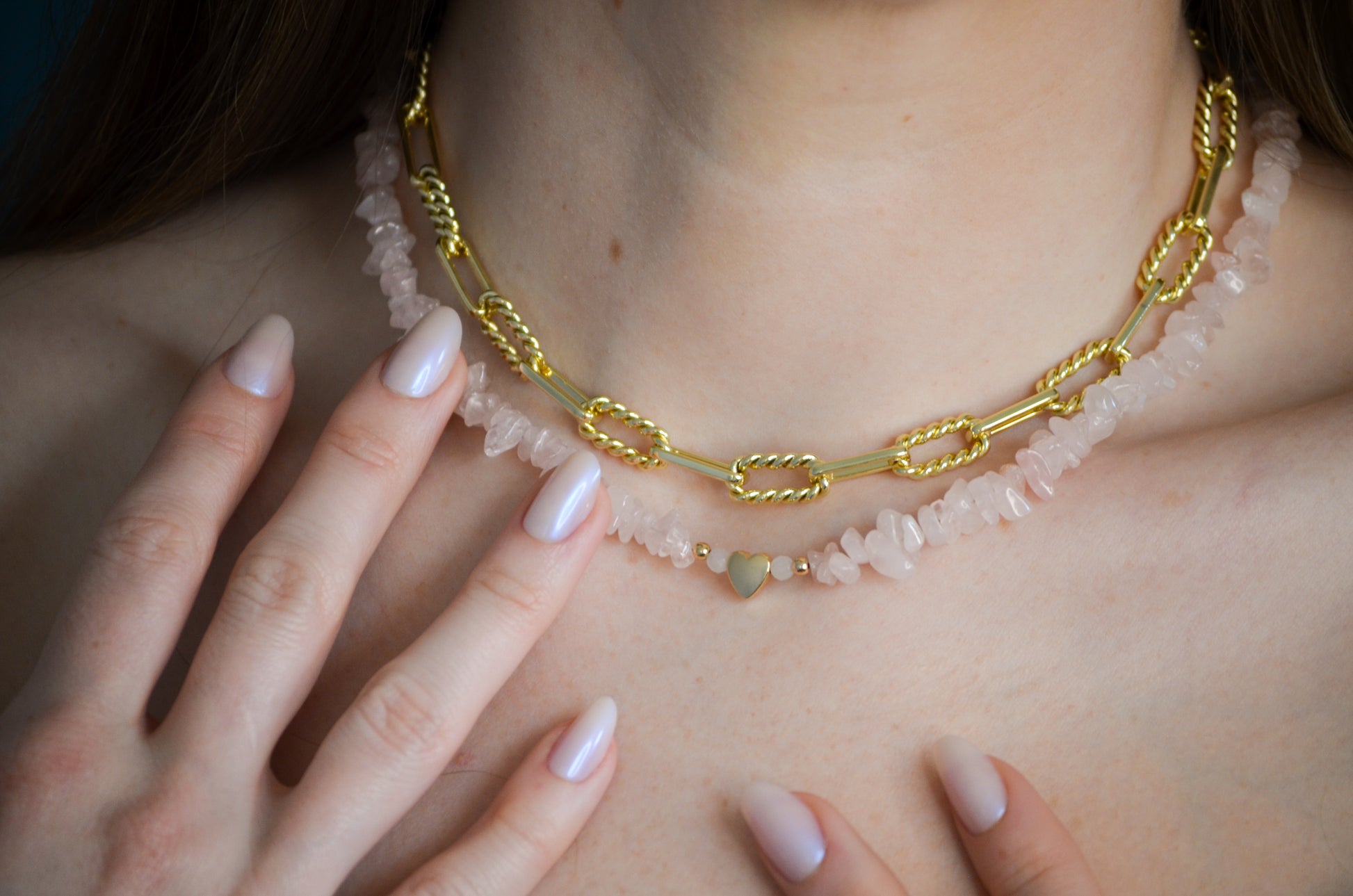 Gold chain necklace with pink stones and gold heart beads worn by a woman with light pink nails