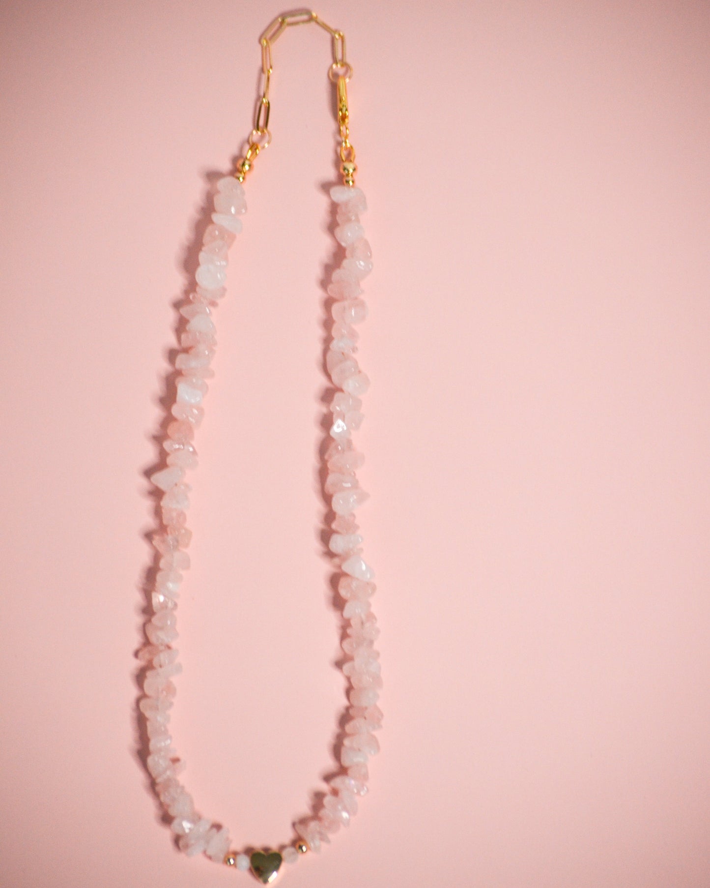 Necklace with pink beads, a gold clasp, and a gold heart charm on a pink background