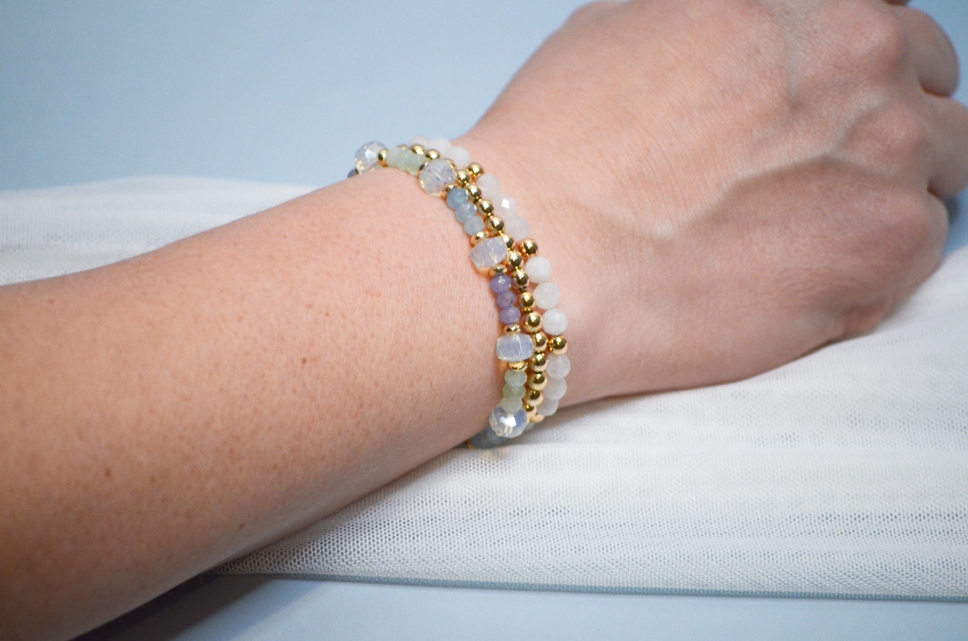 Gold, blue, green, purple, and white beaded bracelet on a wrist against a light blue background