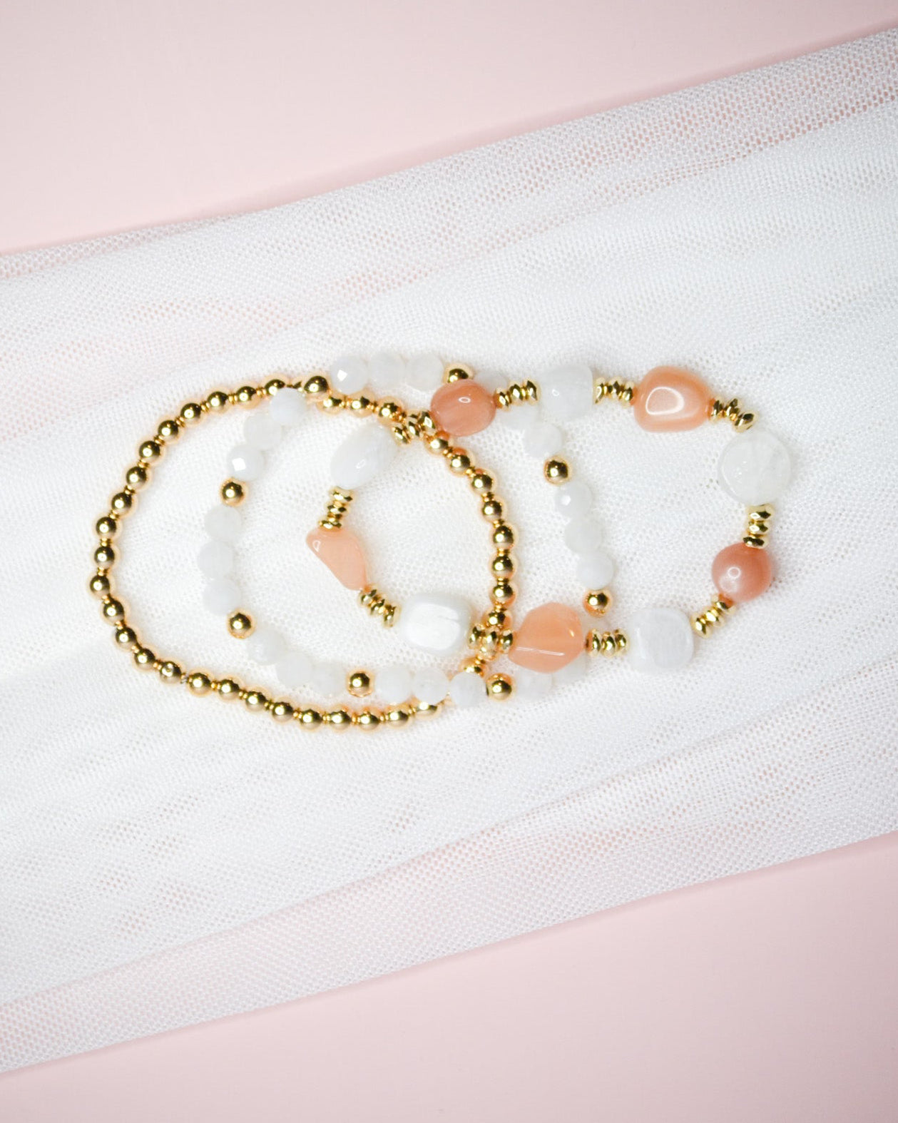 Three bracelets with warm pink, white, and gold beads on a white fabric background