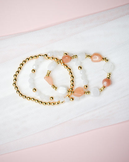 Three bracelets with warm pink, white, and gold beads on a white fabric background