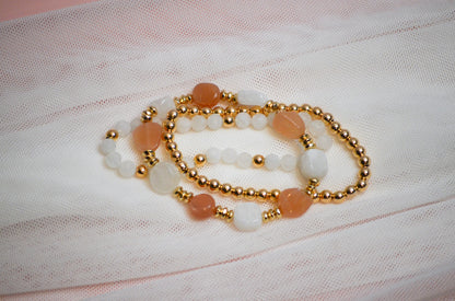 Set of three gold, pink, and white beaded bracelets on a textured fabric background