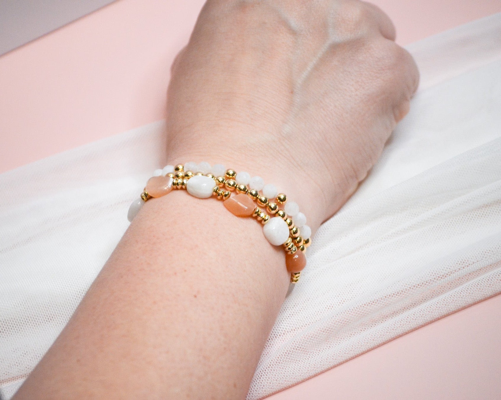 Close-up of a wrist wearing three warm toned and gold beaded bracelets on a light pink background